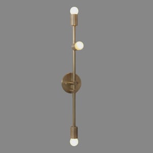 May include: A brass-colored wall sconce with three light bulbs. The fixture has a vertical design with a circular base and a series of cylindrical and spherical elements. The bulbs are illuminated, casting a warm glow. The sconce is designed for indoor lighting.