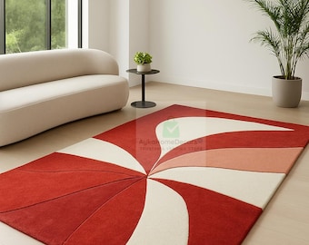 May include: A red, white, and peach-colored area rug with a swirling, geometric design. The rug is rectangular and placed on a light-colored wooden floor. The room also features a white sofa, a small black side table, and a potted plant.