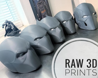 May include: Several gray 3D-printed superhero masks are displayed on a metallic surface. The masks are in various stages of completion, with visible details and a smooth texture. The text "RAW 3D PRINTS" is visible in the image.