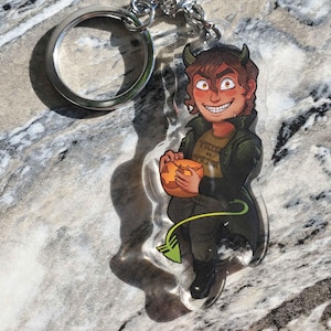 May include: An acrylic keychain featuring a cartoon character with brown hair, small horns, and a green tail. The character wears a dark green coat over a shirt with the text Trick or Treat and holds an orange jack-o'-lantern. The keychain has a silver ring.
