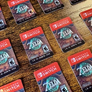 May include: Multiple Nintendo Switch game cartridges for "Zelda" are arranged on a wooden surface. The cartridges have a red border with the Nintendo Switch logo and the game title. The game's artwork is visible on the front.