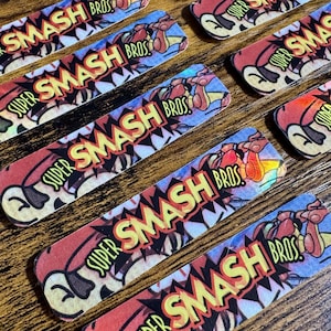 May include: Rectangular stickers with the text "SUPER SMASH BROS" in a comic book style. The stickers feature a red, yellow, and blue color scheme with a cartoon graphic of a boxing glove. The stickers are arranged on a wooden surface.