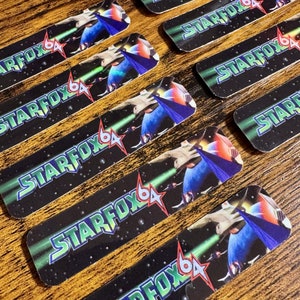 May include: Rectangular black stickers with the text "STARFOX 64" in green and blue, featuring a spaceship and a red star. The stickers are arranged on a wooden surface.