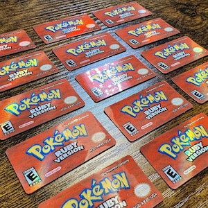 May include: Multiple red Pokémon Ruby Version game cartridges are arranged on a wooden surface. The cartridges feature the Pokémon logo in yellow and blue, with the text "RUBY VERSION" below. The game is rated "E" for Everyone.