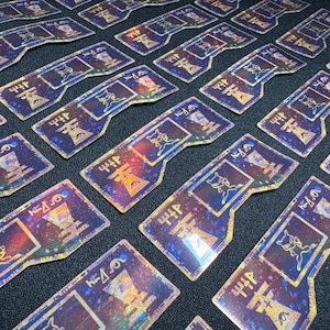 May include: Rectangular, iridescent stickers with a dark blue and purple galaxy design. Each sticker features a stylized cat-like creature, along with symbols and text. The stickers are arranged on a dark surface.