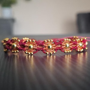 May include: A handmade bracelet featuring a maroon and gold floral design. The bracelet is made of woven maroon thread with gold-colored beads. The bracelet is approximately 7 inches in length and has a pink string closure.