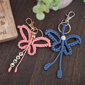 May include: Two macrame butterfly keychains, one pink and one blue, with gold-colored hardware. The pink keychain has the name "LEAH" spelled out in small beads. A wooden plaque with German text is in the background.
