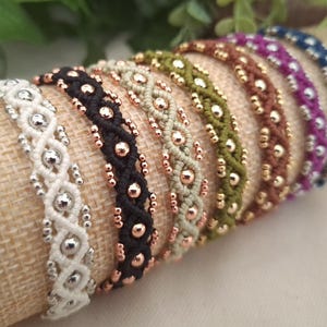 Puede incluir: Una colección de siete pulseras de macramé dispuestas en fila, cada una con un patrón de cordón trenzado integrado con cuentas metálicas. Las pulseras son de color blanco, negro, beige, verde oliva, marrón, morado y azul, con cuentas a juego.