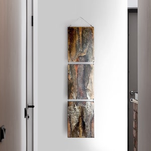 Könnte beinhalten: Ein dreiteiliges abstraktes Kunstwerk mit einer metallischen Oberfläche. Das Kunstwerk besteht aus drei rechteckigen Paneelen, jedes mit einem anderen Design. Die Paneele sind vertikal aufgehängt, wobei das obere Paneel das größte und das untere Paneel das kleinste ist. Das Kunstwerk ist eine Mischung aus braunen, goldenen und schwarzen Farben.