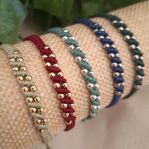 Pode incluir: Cinco pulseiras de cordão trançado dispostas em fila. A primeira é bege com contas douradas, a segunda é vermelho-escura com contas douradas, a terceira é verde-sálvia com contas prateadas, a quarta é azul-marinho com contas prateadas e a quinta é verde-escura com contas prateadas.