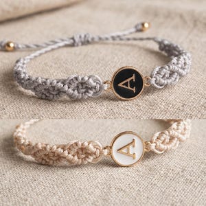 Könnte beinhalten: Zwei Makramee-Armbänder mit einem runden Mittelstück, das einen goldenen Buchstaben A zeigt. Das obere Armband ist grau mit einem schwarzen Emaille-Anhänger, das untere ist beige mit einem weißen Emaille-Anhänger. Beide sind verstellbar.