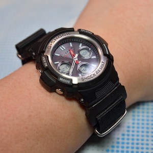 May include: A black and silver G-Shock watch with a black nylon strap. The watch face has a digital display and analogue hands. The watch is on a person's wrist.