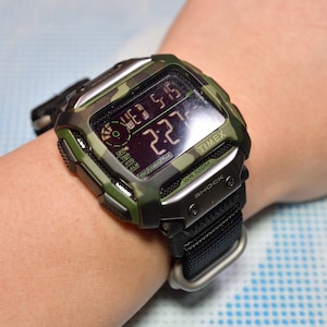 Op de afbeelding: Een digitaal camouflagehorloge in groen en zwart met een zwarte nylon band. De wijzerplaat van het horloge toont de tijd, datum en dag van de week. Het horloge is waterdicht tot 100 meter en heeft een schokbestendige functie. Het horlogemerk is Timex.