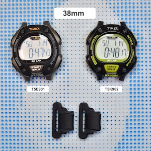 May include: Two black digital watches with Ironman Triathlon branding. The watch on the left has a grey face and the watch on the right has a green face. Both watches have a black band and are labelled with the model number. The watches are on a blue and white patterned surface.
