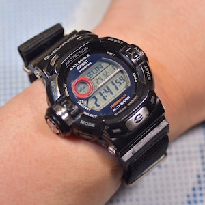 Op de afbeelding: Een zwart Casio G-Shock horloge met een zwarte nylon band. De wijzerplaat van het horloge toont de tijd, datum en dag van de week. Het horloge heeft ook een digitale hoogtemeter en een kompas.