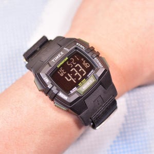 Puede incluir: Reloj digital Timex Expedition negro que muestra la hora 4:33. El reloj tiene una correa negra y detalles en verde. El reloj está en una muñeca.