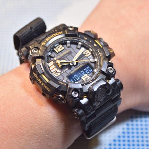 May include: A black and gold G-Shock watch with a digital display and a black resin band. The watch has a large, round face with a black and gold bezel. The watch is on a person's wrist.