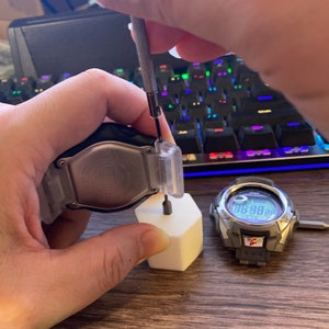 May include: A person is using a small screwdriver to remove the back of a silver watch. The watch is sitting on a white hexagonal stand. A second watch with a black face and a digital display is visible in the background.