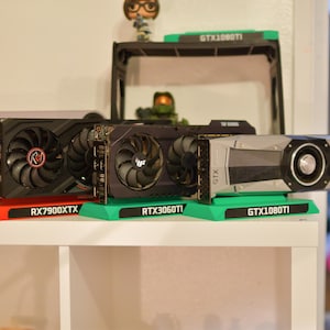 May include: Three graphics cards displayed on individual stands. The left card is black with the text RX7900XTX. The middle card is black with the text RTX3060TI. The right card is silver and black with the text GTX1080TI. Each stand features the model name.