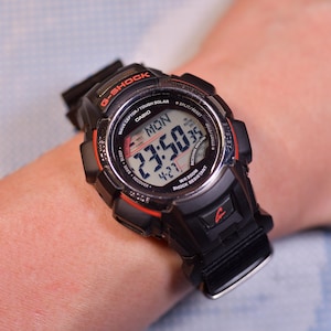 May include: Black G-Shock digital watch with red accents, displaying the time as 23:50:35 on a Monday. The watch features a digital display and is shock-resistant.