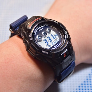 Puede incluir: Reloj digital negro con una correa de tela azul, que muestra la hora 3:31:24. La esfera del reloj tiene el texto "G-SHOCK" y "CASIO". El reloj está en una muñeca.