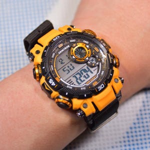 May include: A black and yellow digital wristwatch with a black strap. The watch face has a digital display showing the time, date, and other features. The watch has a gold-coloured bezel and buttons.
