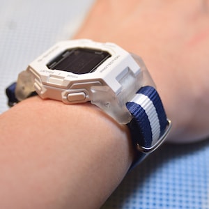 Könnte beinhalten: Weiße Digitaluhr mit transparentem Schutzgehäuse und einem marineblau-weiß gestreiften Armband. Die Uhr hat ein rechteckiges Zifferblatt mit dunkler Anzeige und weißen Knöpfen. Das Armband wird mit einer silbernen Schnalle befestigt.