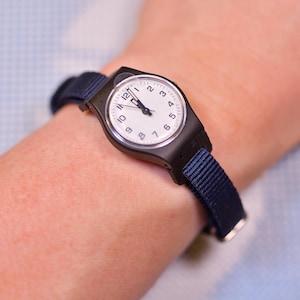 Könnte beinhalten: Eine schwarze Armbanduhr mit weißem Zifferblatt und schwarzen Ziffern. Die Uhr hat ein marineblaues Stoffarmband. Die Uhr wird am Handgelenk getragen.