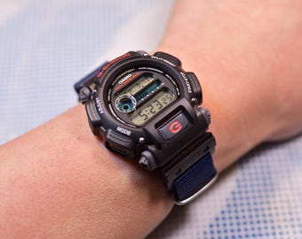 May include: A black Casio G-Shock watch with a digital display showing the time as 5:23:25. The watch has a black band with blue stitching and a red G-Shock logo.