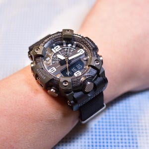 May include: A black and green G-Shock watch with a carbon fiber face and a black nylon strap. The watch has a digital display and a large analog face with white hands and markers. The watch is on a person's wrist.