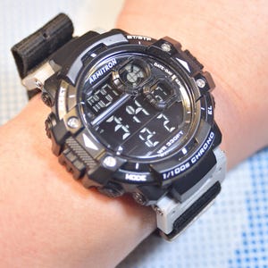 May include: A black Armitron digital watch with a black and gray nylon strap. The watch face has a black background with white numbers and a white date window. The watch is water resistant to 330 feet.