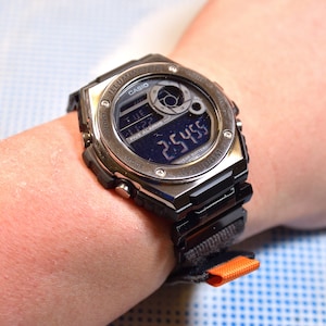 May include: A silver and black Casio watch with a digital display showing the time and date. The watch has a black nylon strap with an orange accent.