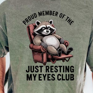 Op de afbeelding: Olijfgroen t-shirt met een cartoon wasbeer in een rode fauteuil. De tekst luidt "Proud Member of the Just Resting My Eyes Club". De wasbeer heeft een zwart masker en glimlacht.