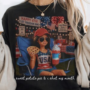 May include: A black t-shirt featuring a graphic of a person in a red cap and sunglasses sitting in a blue chair, holding a pie and a red, white, and blue drink. The text reads "HAPPY 4TH! LET FREEDOM RING" and "sweet potato pie & i shut my mouth".