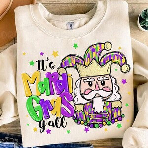 Puede incluir: Una sudadera blanca con el texto "It's Mardi Gras y'all" en letras moradas, verdes y amarillas. Un bufón de dibujos animados con una corona y ropa con estampado morado, verde y dorado se encuentra en el centro del diseño.