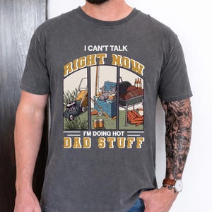 Può includere: T-shirt grigio scuro con la scritta "I CAN'T TALK RIGHT NOW I'M DOING HOT DAD STUFF". La grafica raffigura un tosaerba, attrezzatura da pesca e un barbecue. La maglietta è realizzata in materiale morbido.