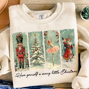 Puede incluir: Una sudadera de color crema con un diseño navideño de estilo vintage. El diseño presenta un cascanueces, un árbol de Navidad, una bailarina y un rey ratón. El texto "Have yourself a merry little Christmas" está impreso debajo del diseño.