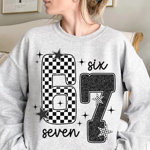 Może przedstawiać: Jasnoszara bluza z numerem "67" w szachownicę i wzór w cętki. Słowo "six" znajduje się nad numerem, a "seven" pod nim. Czarne gwiazdki i krzyżyki dopełniają projekt.