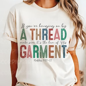 May include: A white t-shirt with a colorful graphic that reads "If you're hanging on by a thread make sure it's the hem of His garment" with the scripture reference "Matthew 9:20-22" underneath.