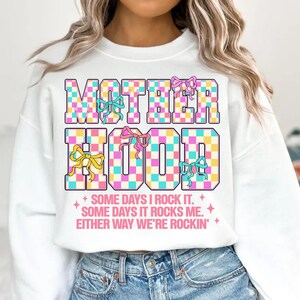 May include: White sweatshirt with a checkered design that says "MOTHERHOOD" in pink, blue, and yellow. The text below says "SOME DAYS I ROCK IT. SOME DAYS IT ROCKS ME. EITHER WAY WE'RE ROCKIN'."