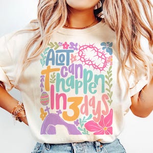May include: A light beige t-shirt with a colorful graphic design that says "A lot can happen in 3 days". The design features a crown of thorns, an Easter egg, a cross, and flowers.