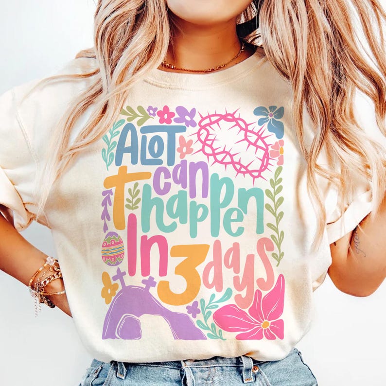 May include: A light beige t-shirt with a colorful graphic design that says "A lot can happen in 3 days". The design features a crown of thorns, an Easter egg, a cross, and flowers.