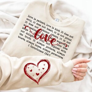 Puede incluir: Sudadera beige con un corazón rojo de purpurina y una cita de 1 Corintios 13:4-7 sobre el amor. La cita dice: "El amor es paciente, el amor es bondadoso. No tiene envidia, no se jacta, no se enorgullece. No busca lo suyo, no se irrita, no guarda rencor. El amor no se alegra de la injusticia, sino que se alegra con la verdad. Todo lo disculpa, todo lo cree, todo lo espera, todo lo soporta. 1 Corintios 13:4-7"