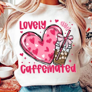 Puede incluir: Sudadera blanca con un corazón rosa y una taza de café rosa y marrón con un lazo rosa y una pajita. El texto "Lovely and Caffeinated" está impreso en la camiseta.
