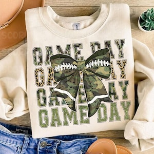 May include: A beige sweatshirt with the text "GAME DAY" repeated four times in a stacked design. The text is in a green and black camouflage pattern with a large green and black camouflage bow with white accents in the center.