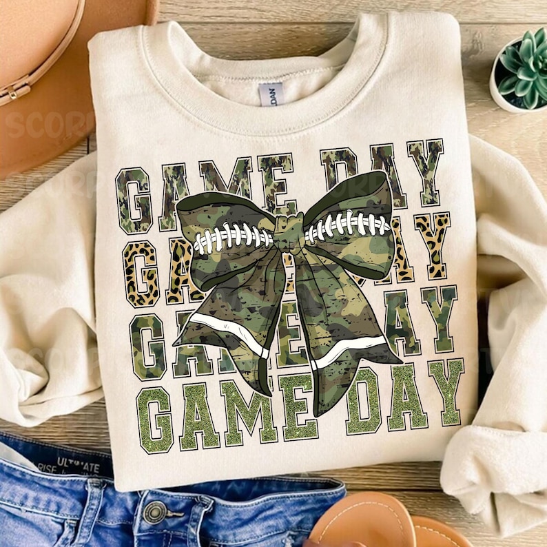 May include: A beige sweatshirt with the text "GAME DAY" repeated four times in a stacked design. The text is in a green and black camouflage pattern with a large green and black camouflage bow with white accents in the center.