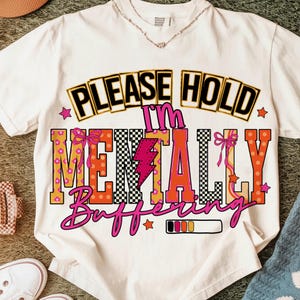 May include: White t-shirt with the text "PLEASE HOLD I'M MENTALLY Buffering" in a colorful, retro design. The design includes a lightning bolt, bows, stars, and a loading bar. The shirt is a casual wear item.