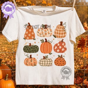 Pode incluir: Uma t-shirt de cor creme com uma impressão gráfica de nove abóboras com vários padrões, incluindo flores, xadrez, padrão de leopardo e bolinhas. O design inclui pequenas estrelas e o texto Scorpiosartvn num emblema circular.