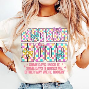 May include: A white t-shirt with the text "MOTHERHOOD" in pink, blue and yellow checkered letters with pink bows. The text "SOME DAYS I ROCK IT. SOME DAYS IT ROCKS ME. EITHER WAY WE'RE ROCKIN'" is printed below the main text.