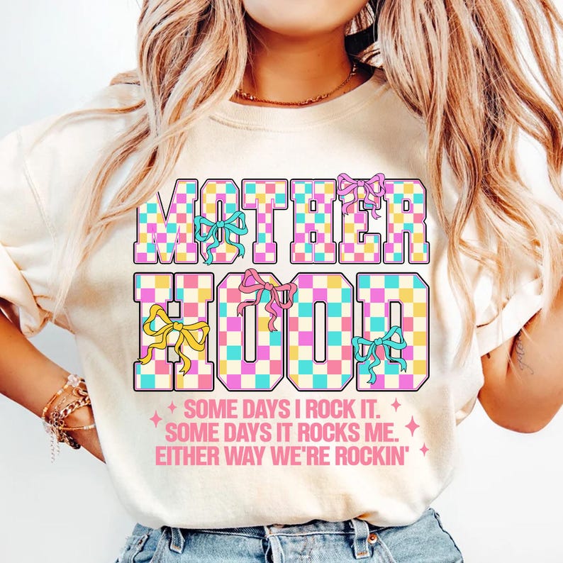 May include: A white t-shirt with the text "MOTHERHOOD" in pink, blue and yellow checkered letters with pink bows. The text "SOME DAYS I ROCK IT. SOME DAYS IT ROCKS ME. EITHER WAY WE'RE ROCKIN'" is printed below the main text.